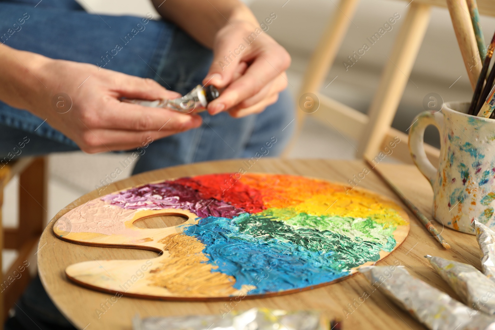 Photo of Man opening tube of paint at wooden table with palette indoors, closeup