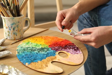 Photo of Man opening tube of paint at wooden table with palette indoors, closeup