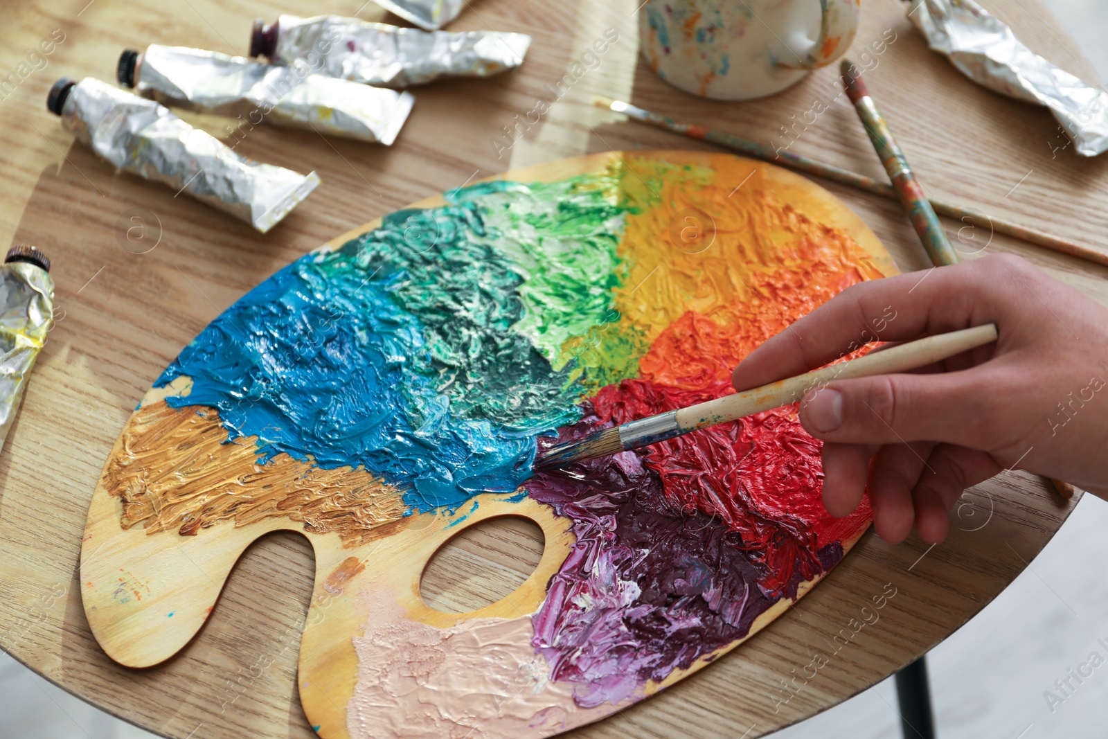 Photo of Man with paintbrush mixing paints on palette at wooden table, closeup