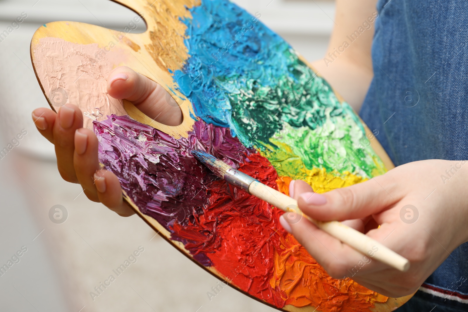 Photo of Woman with paintbrush mixing paints on palette against blurred background, closeup