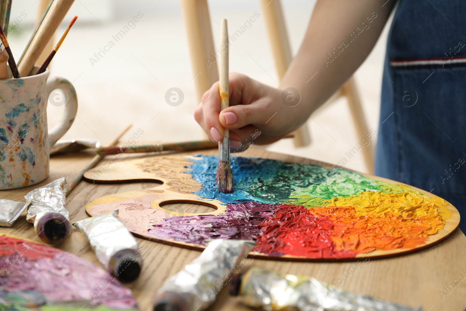 Photo of Woman with paintbrush mixing paints on palette at wooden table indoors, closeup