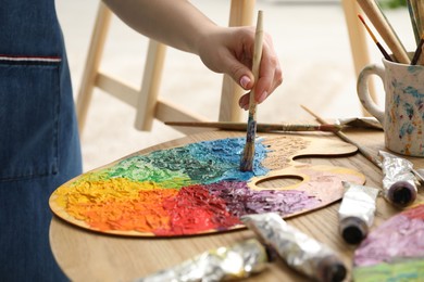 Photo of Woman with paintbrush mixing paints on palette at wooden table indoors, closeup