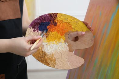 Photo of Woman with paintbrush mixing paints on palette near picture indoors, closeup