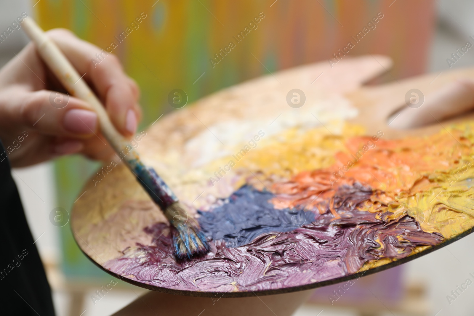 Photo of Woman with paintbrush mixing paints on palette against blurred background, closeup