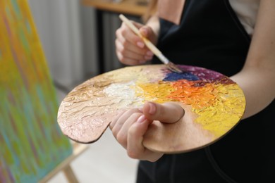 Photo of Woman with paintbrush mixing paints on palette near picture indoors, closeup