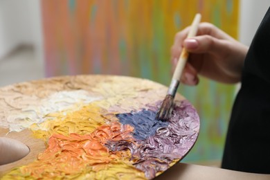 Photo of Woman with paintbrush mixing paints on palette against blurred background, closeup