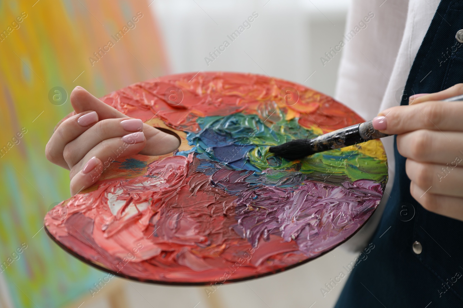 Photo of Woman with paintbrush mixing paints on palette against blurred background, closeup