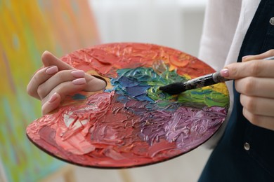 Woman with paintbrush mixing paints on palette against blurred background, closeup