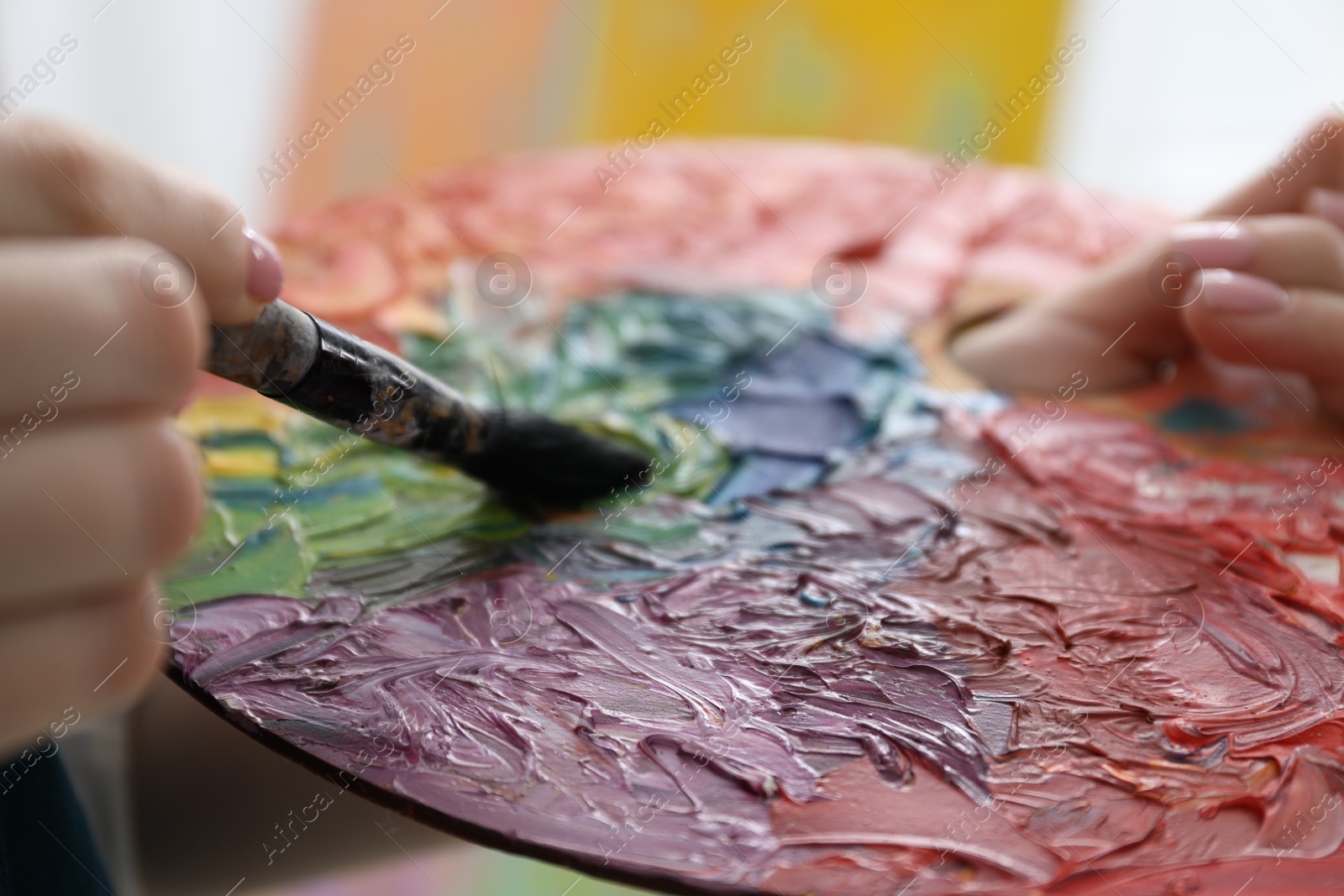 Photo of Woman with paintbrush mixing paints on palette against blurred background, closeup