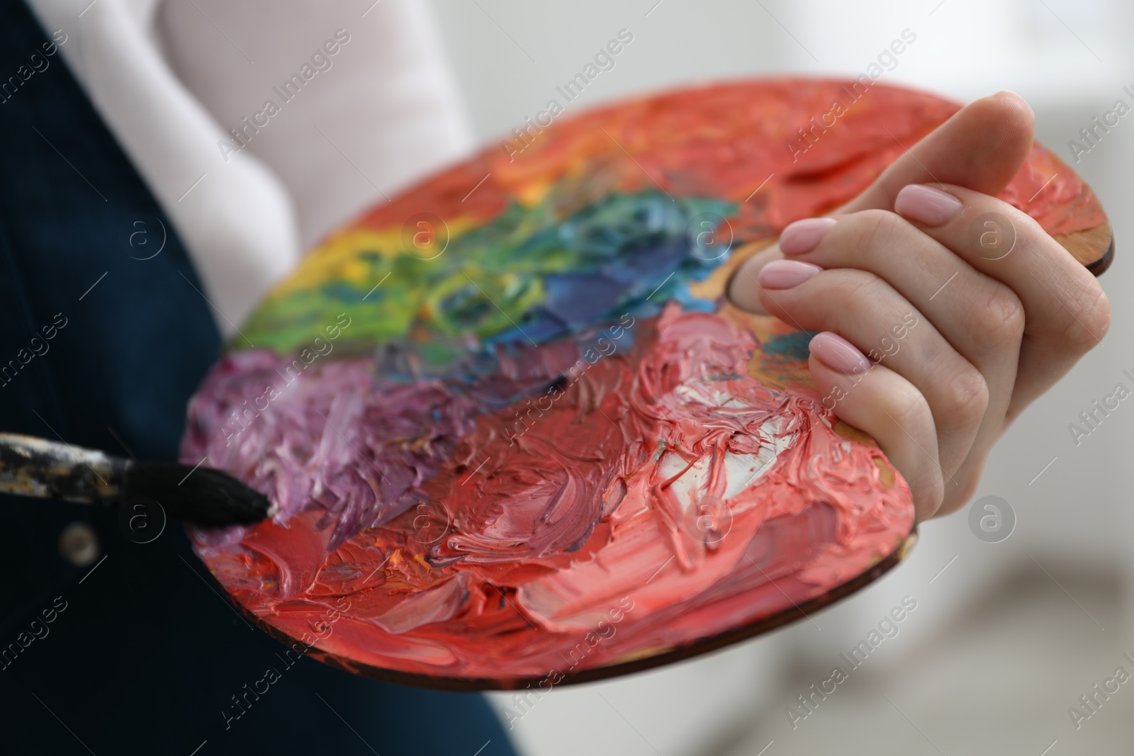 Photo of Woman with paintbrush mixing paints on palette against blurred background, closeup