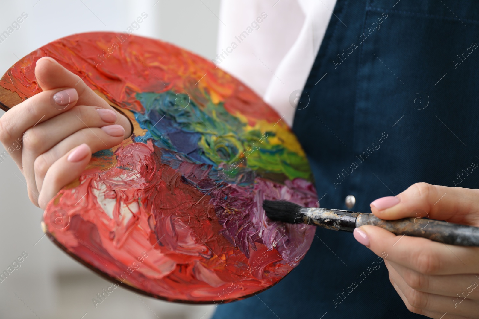 Photo of Woman with paintbrush mixing paints on palette against blurred background, closeup