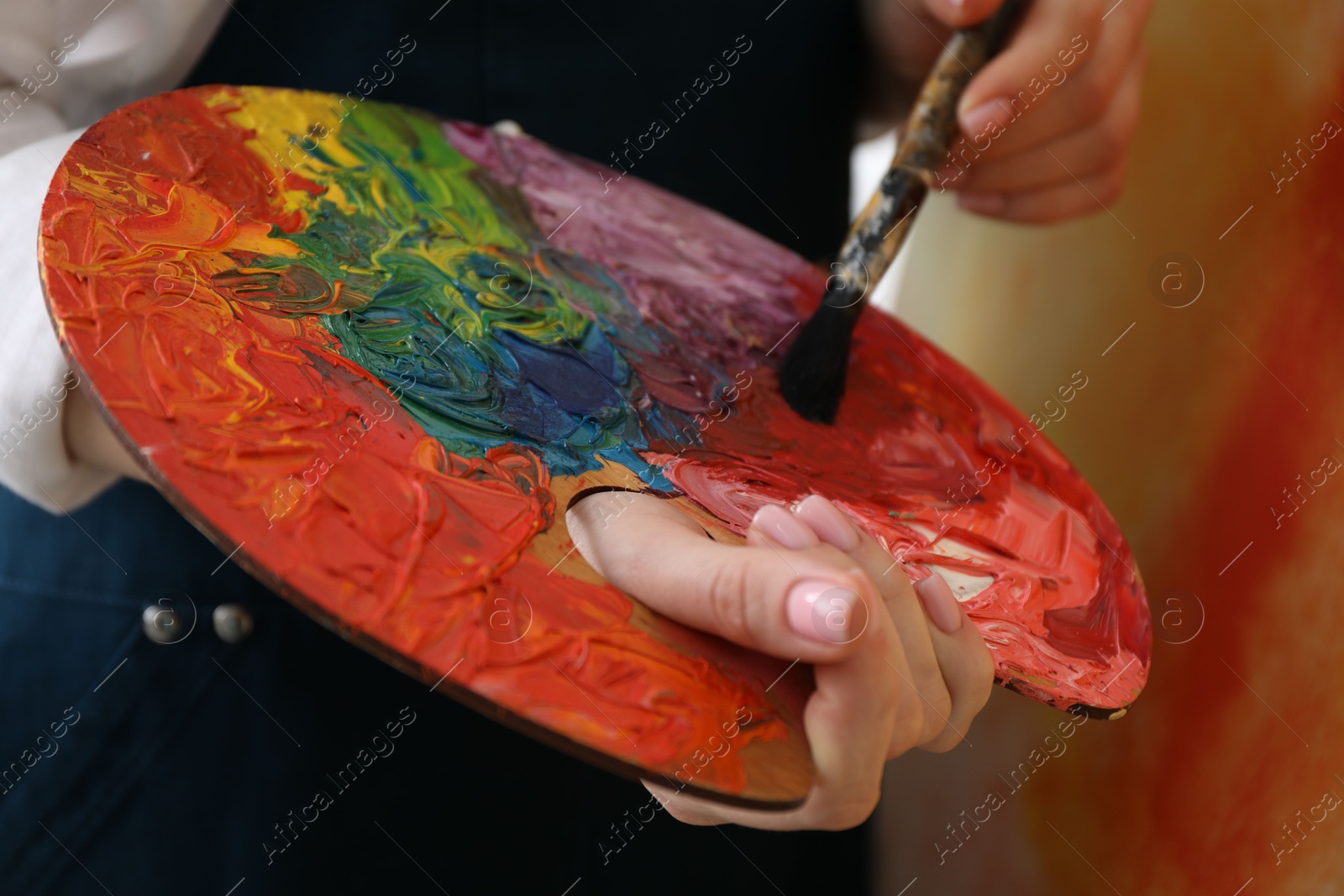 Photo of Woman with paintbrush mixing paints on palette against blurred background, closeup