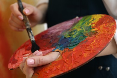 Photo of Woman with paintbrush mixing paints on palette against blurred background, closeup