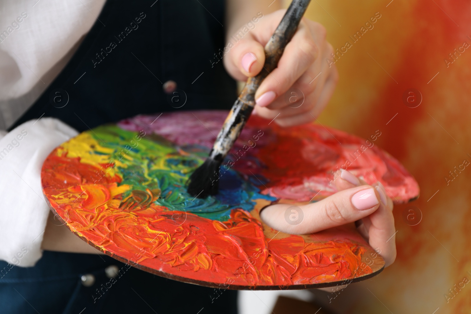 Photo of Woman with paintbrush mixing paints on palette against blurred background, closeup