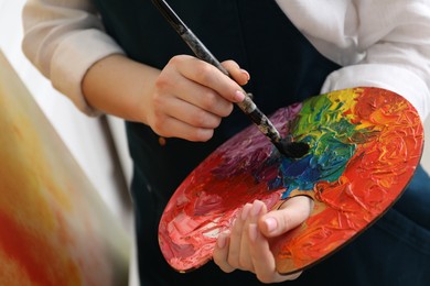 Photo of Woman with paintbrush mixing paints on palette indoors, closeup