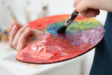 Photo of Woman with paintbrush mixing paints on palette against blurred background, closeup