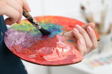 Photo of Woman with paintbrush mixing paints on palette against blurred background, closeup