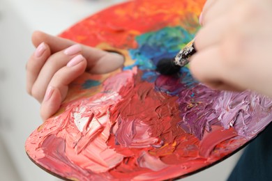 Woman with paintbrush mixing paints on palette against blurred background, closeup