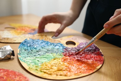 Woman with paintbrush mixing paints on palette at wooden table indoors, closeup