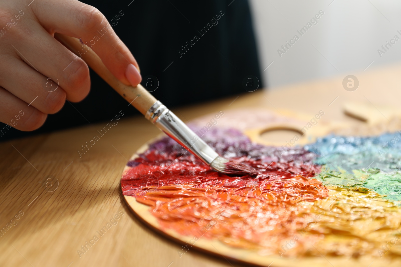 Photo of Woman with paintbrush mixing paints on palette at wooden table indoors, closeup