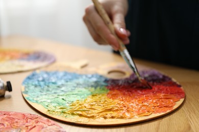 Photo of Woman with paintbrush mixing paints on palette at wooden table indoors, closeup