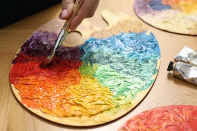 Photo of Woman with paintbrush mixing paints on palette at wooden table, closeup