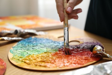 Woman with paintbrush mixing paints on palette at wooden table, closeup