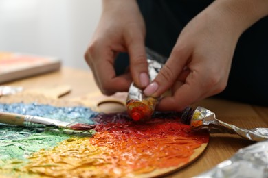 Photo of Woman squeezing paint from tube onto on palette at wooden table, closeup