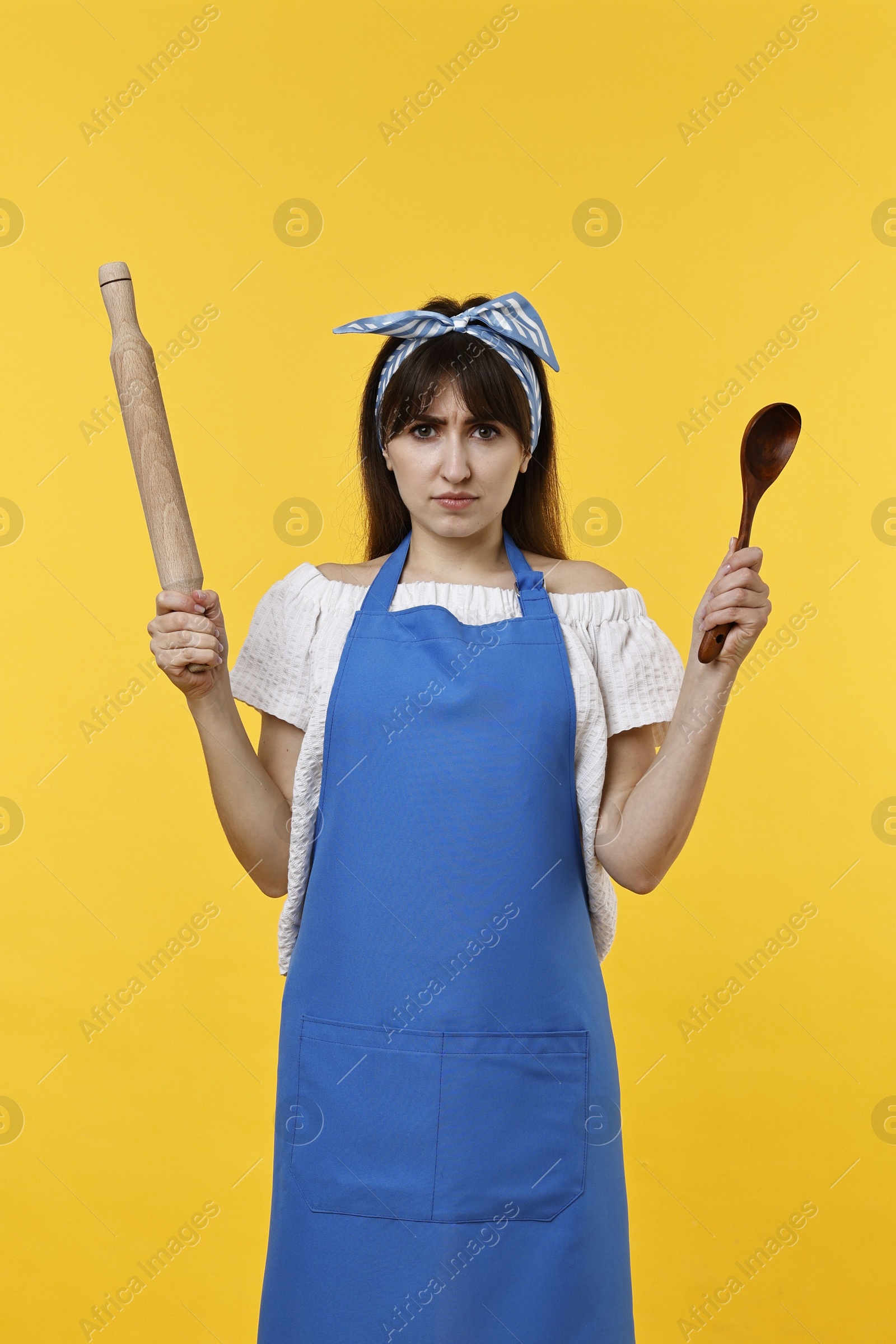 Photo of Angry woman with rolling pin and spoon on yellow background