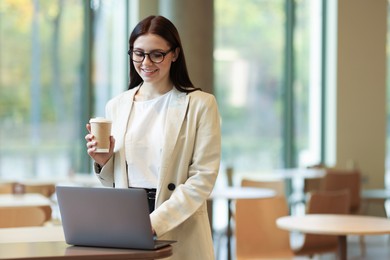 Photo of Woman in stylish formal suit with coffee working on laptop indoors