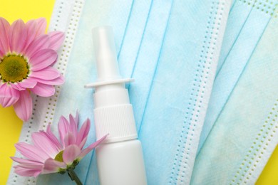 Photo of Allergy treatment. Nasal spray, protective masks and flowers on yellow background, top view