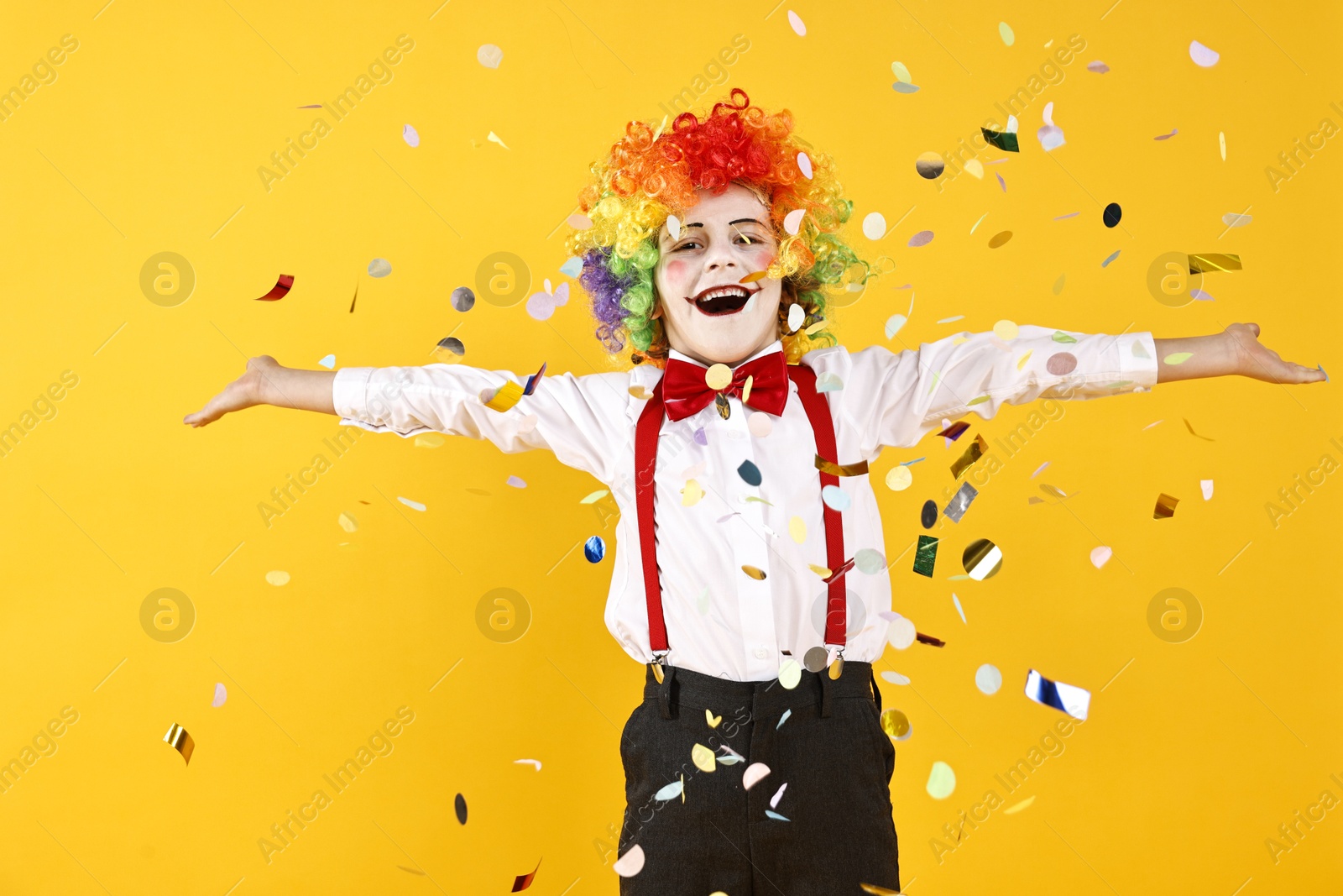 Photo of Happy little boy dressed like clown and flying confetti on yellow background. Surprise party