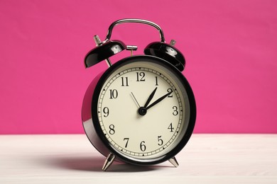 Photo of One black alarm clock on white wooden table against bright pink background