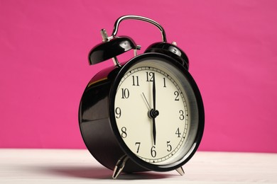 Photo of One black alarm clock on white wooden table against bright pink background