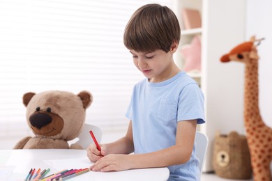 Photo of Cute boy drawing near teddy bear at white table in kindergarten