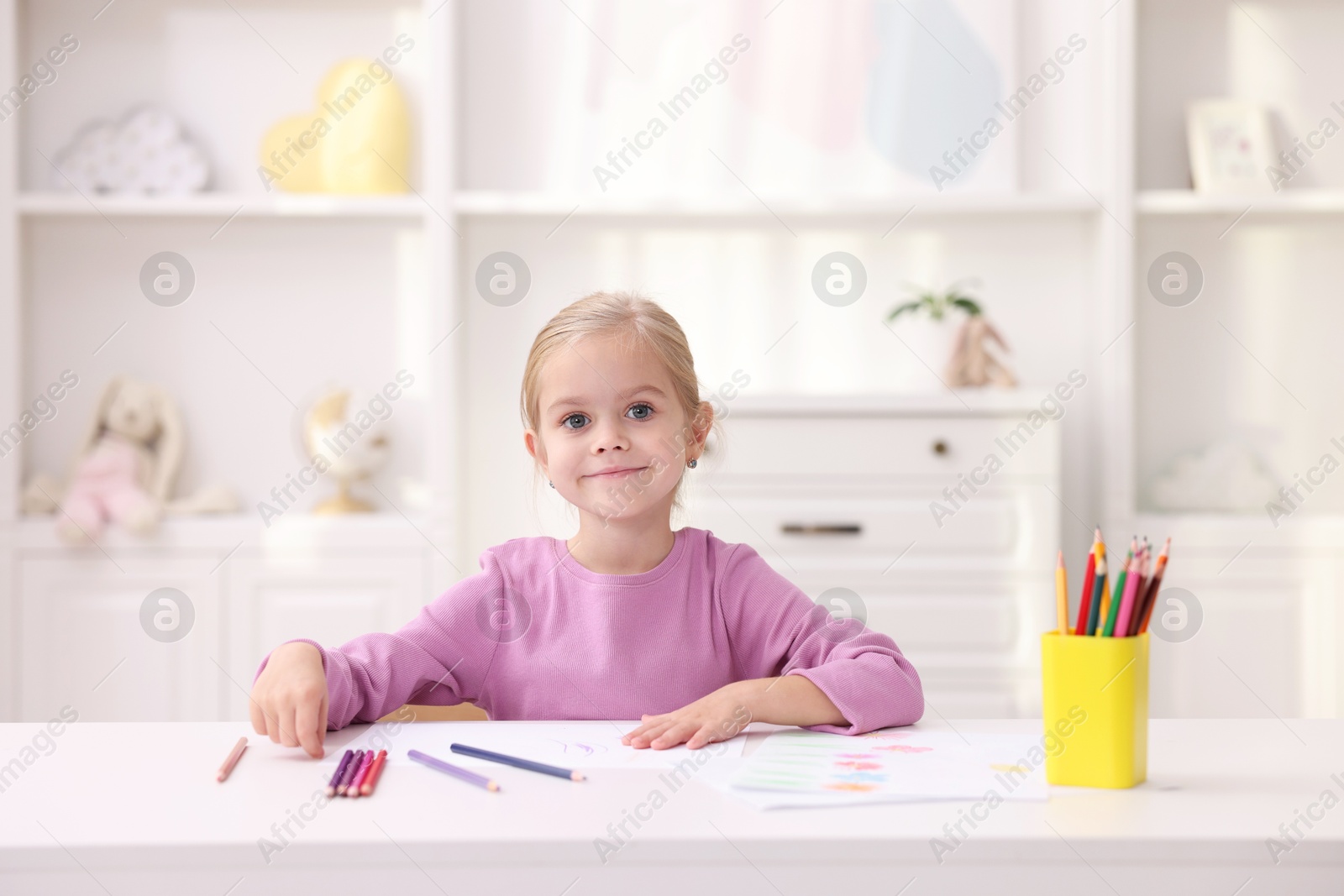 Photo of Cute girl at white table with drawing and pencils in kindergarten