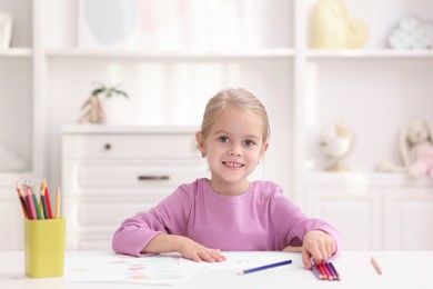 Photo of Happy girl at white table with drawing and pencils in kindergarten