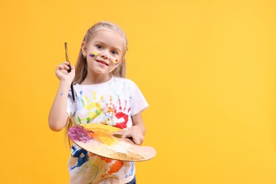 Photo of Smiling girl with brush and palette smeared in paint on orange background. Space for text