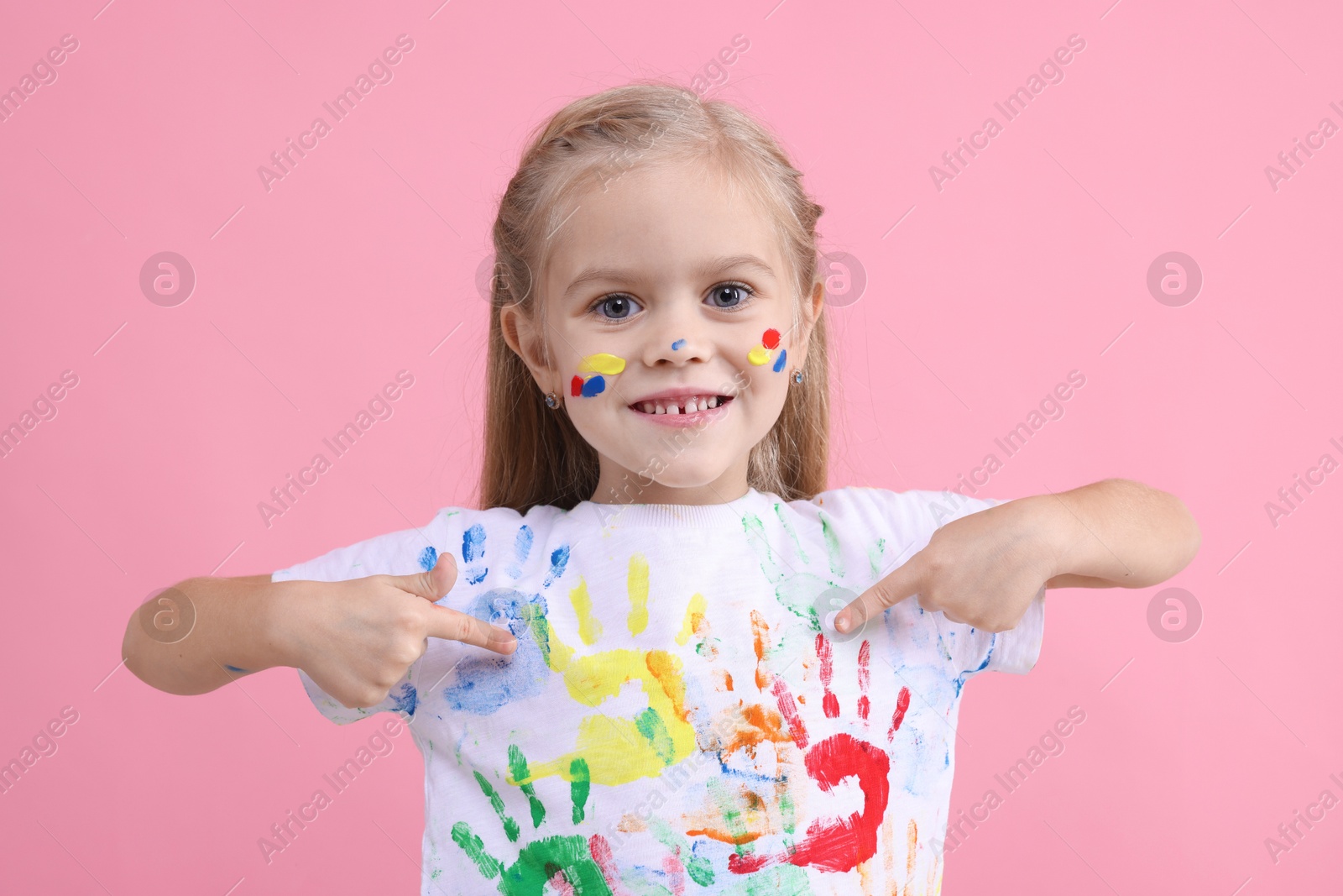 Photo of Portrait of smiling girl pointing at her t-shirt with handprints drawings on pink background