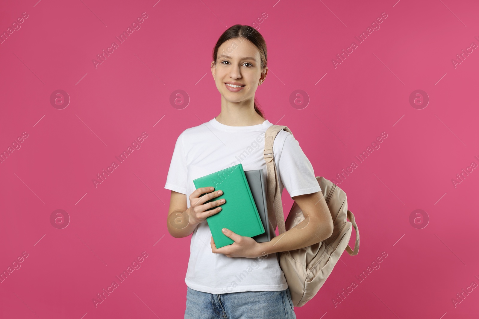 Photo of Portrait of smiling teenage girl with books and backpack on pink background