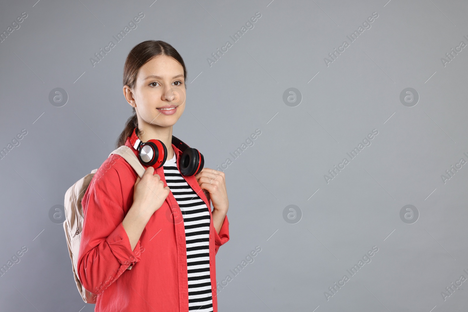 Photo of Portrait of smiling teenage girl with backpack and headphones on grey background. Space for text