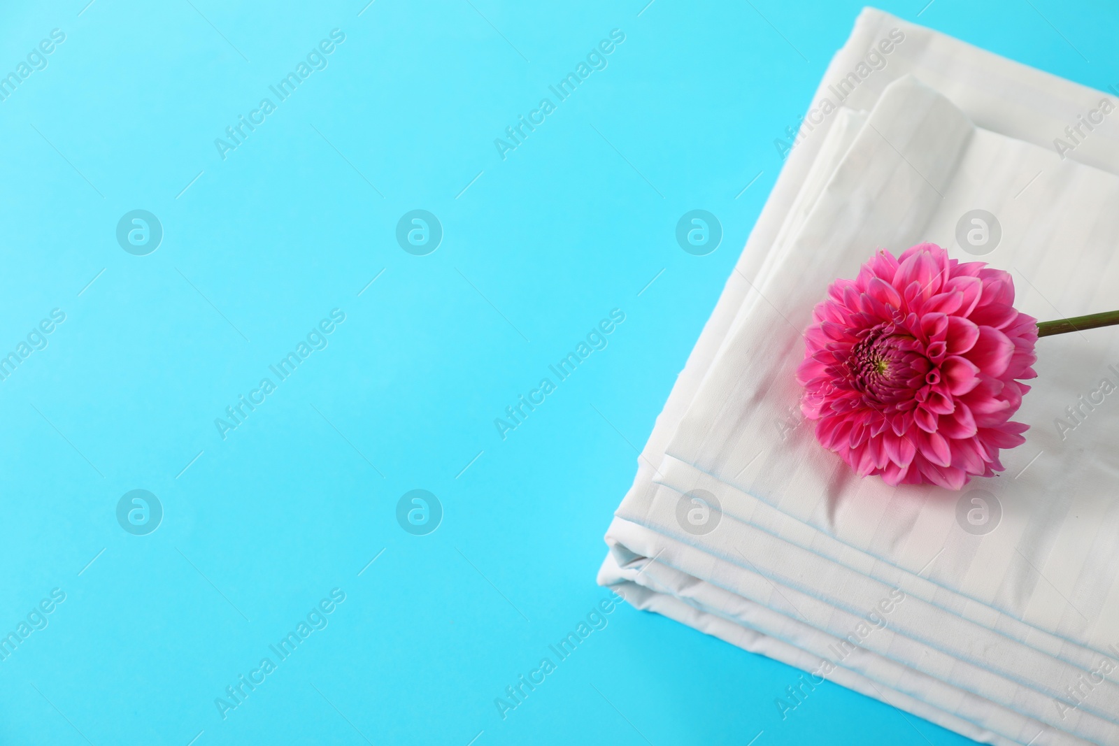 Photo of Stacked clean bed linens and pink flower on light blue background, top view. Space for text