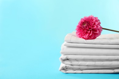 Photo of Stacked clean bed linens and pink flower on light blue background, space for text