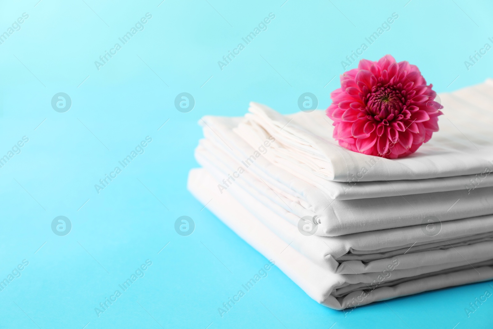 Photo of Stacked clean bed linens and pink flower on light blue background, space for text