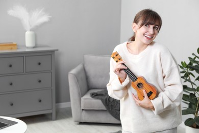 Photo of Happy young woman playing ukulele at home, space for text