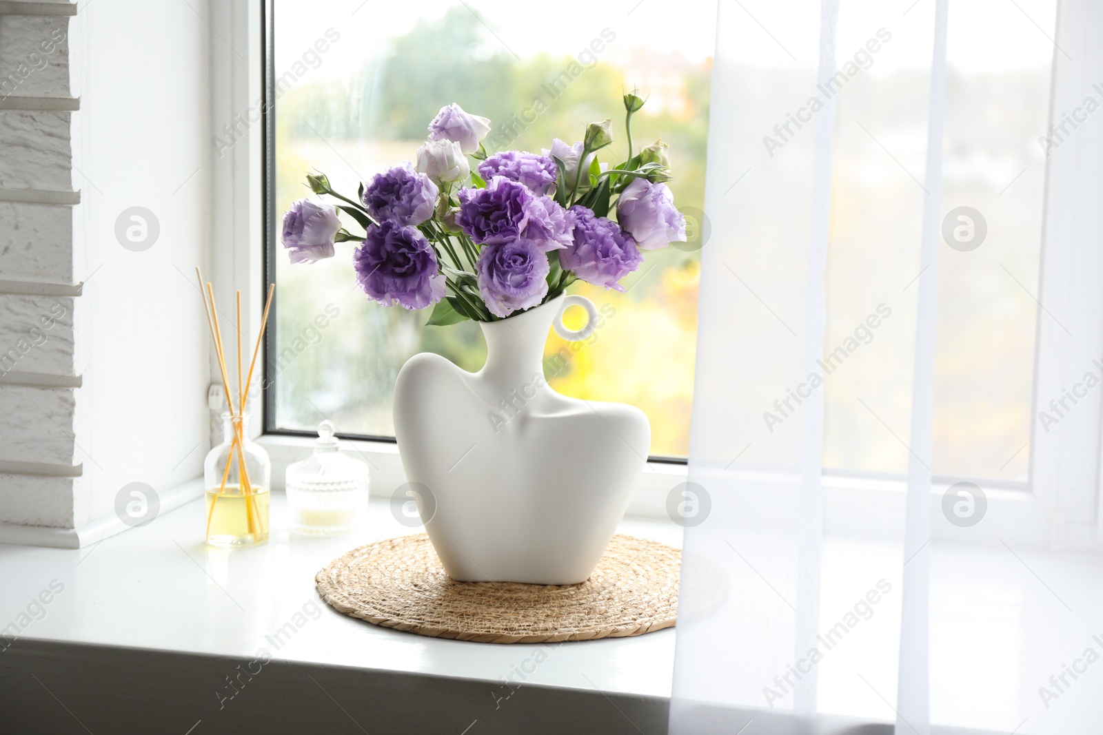 Photo of Stylish vase with beautiful flowers, candle and reed diffuser near window at home