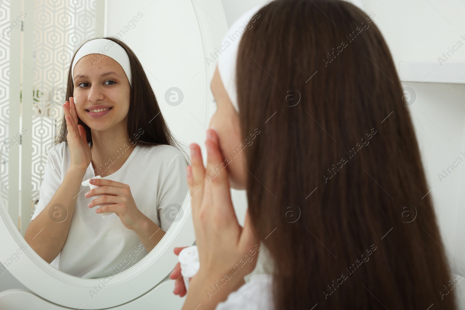 Photo of Teenage girl applying cream onto face near mirror indoors. Acne treatment