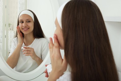 Photo of Teenage girl applying cream onto face near mirror indoors. Acne treatment