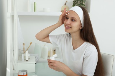 Photo of Teenage girl applying cream onto face near mirror indoors. Acne treatment