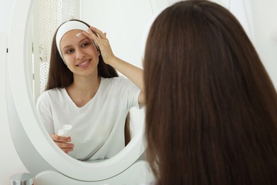 Photo of Teenage girl applying cream onto face near mirror indoors. Acne treatment